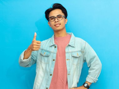 Sıradan giyinmiş genç bir Asyalı kendine güvenen bir jest yapıyor, olumlu bir ifadeyle gülümsüyor. Mavi arka planda izole edilmiş, reklam ve promosyon için mükemmel..