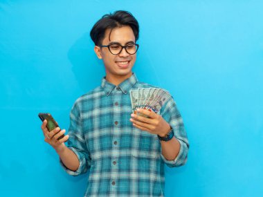 Mavi tişörtlü heyecanlı Asyalı adam elinde cep telefonu ve parayla mavi arka planda izole edilmiş mutlu bir yüz ifadesi gösteriyor..