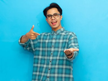 Günlük giysiler içinde genç Asyalı bir adam heyecanlı bir ifadeyle ürün sunumu için boş bir avuç içiyle işaret ediyor. Reklamcılık ya da promosyonlar için mükemmel mavi arka plan, izole edilmiş.