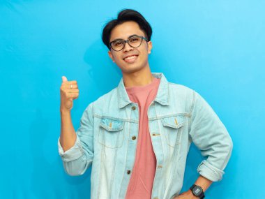 Kendine güvenen genç Asyalı bir adam başparmağıyla kenarı gösterirken poz veriyor, mavi arka planda pozitif bir ifadeyle gülümsüyor, reklam veya promosyon için mükemmel..