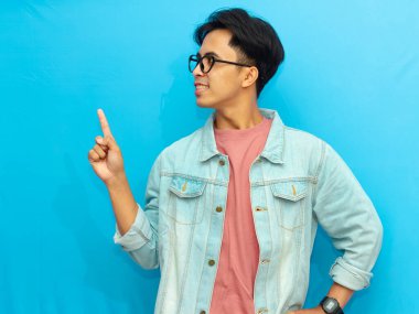 Tek parmağıyla yukarı doğru bakan, mavi arka planda izole edilmiş, Asyalı genç bir adam. Reklam, promosyon ve yaratıcı içerik için mükemmel bir imaj..