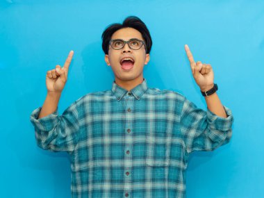 Mavi gömlekli gülümseyen Asyalı adam mavi arka planda poz veriyor ve işaret ediyor. Fikir, ilham veya başarı kavramları için ideal..