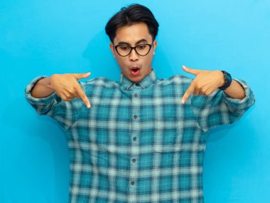 Mavi tişörtlü Asyalı adam gülümsüyor ve mavi arka plana doğru işaret ediyor, neşeli bir ifadeyle pozitif bir jest yapıyor..