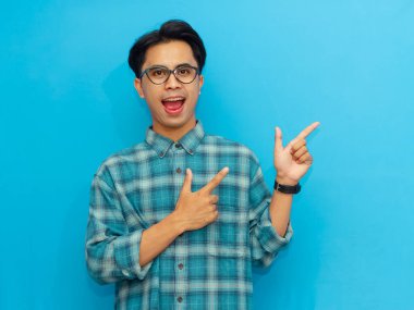 Mavi tişörtlü gülümseyen Asyalı adam mavi arka planı işaret ederek pozitifliği ve dostça iletişimi ifade ediyor..