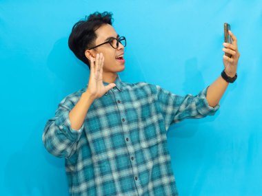 Mavi tişörtlü genç Asyalı bir adam elinde akıllı telefon tutarken gülümsüyor ve mavi arka planda selfie çekiyormuş gibi poz veriyor..