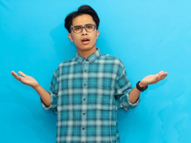 Mavi gömlekli Asyalı bir adam şok olmuş bir ifadeyle iki elini de mavi arka planda karmaşa içinde kaldırıyor.