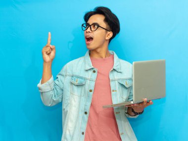 Yakışıklı Asyalı erkek öğrenci elinde dizüstü bilgisayarla beyaz bir arkaplanda izole edilmiş hayret verici bir ifadeyi işaret ediyor..