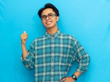 Asian man in a blue casual shirt smiling and giving a thumbs-up gesture while pointing to the side against a blue background. clipart