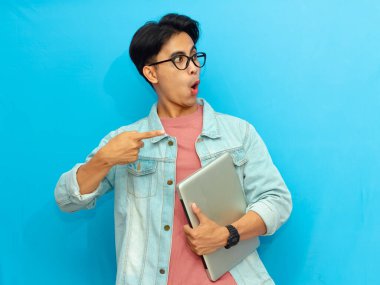 Yakışıklı Asyalı bir erkek öğrenci dizüstü bilgisayar tutuyor ve beyaz bir arkaplanda izole edilmiş hayret verici bir ifadeyle yüzünü gösteriyor..