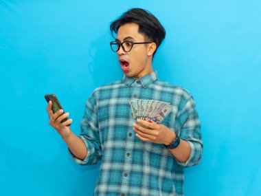 Mavi gömlekli Asyalı adam elinde para tutuyordu ve telefonuna bakıyordu. Mavi arka planda izole edilmiş komik bir şok ifadesi vardı..