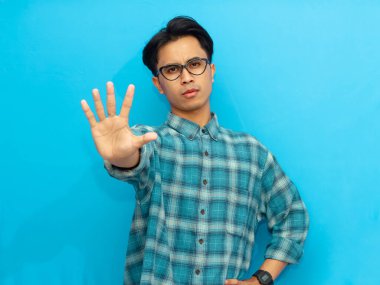 Young Asian man in casual clothing posing with a stop hand gesture on an isolated blue background. Perfect for advertisement, campaign, or awareness promotion. clipart