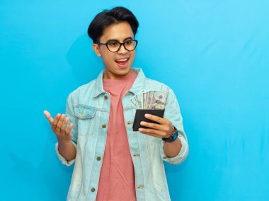 Kot ceketli heyecanlı Asyalı adam mavi arka planda sürpriz bir yüz ifadesi gösteren para dolu bir cüzdanla poz veriyor..
