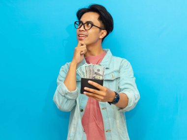 Kot ceketli Asyalı adam elinde para dolu bir cüzdanla gülümsüyor mavi arka planda izole edilmiş düşünceli bir ifadeyle yukarı bakıyor..