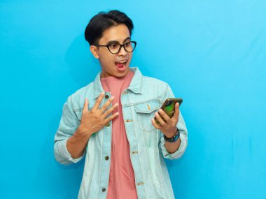 Kot ceketli Asyalı adam elinde harika bir ifadeyle akıllı telefon tutuyor, mavi arka planda izole edilmiş, cep telefonunda gördüklerinden şok olmuş..