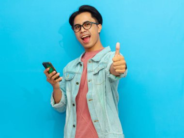 Kot ceketli gülümseyen Asyalı bir adam cep telefonu tutuyor ve mavi arka planda izole bir şekilde kameraya işaret veriyor..