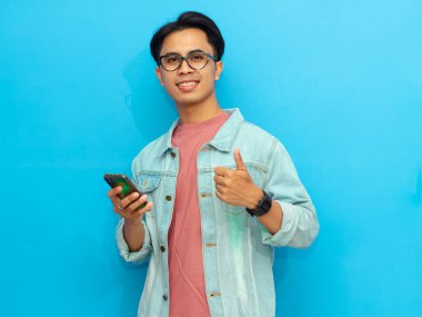 Kot ceketli gülümseyen Asyalı bir adam cep telefonu tutuyor ve mavi arka planda izole bir şekilde kameraya işaret veriyor..