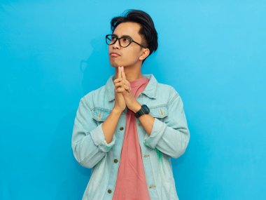 Kot ceket giymiş Asyalı bir adam mavi bir arka planda izole edilmiş, düşünceli bakışlar atıyor..