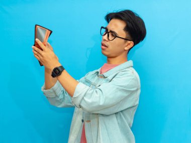 Kot ceketli zavallı Asyalı adam mavi arka planda soyutlanmış, üzgün bir ifadeyle boş bir cüzdan taşıyor..