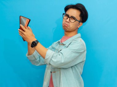 Poor Asian man in a denim jacket holding an empty wallet with a sad expression, isolated on a blue background. clipart