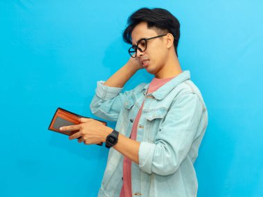 Kot ceketli zavallı Asyalı adam mavi arka planda soyutlanmış, üzgün bir ifadeyle boş bir cüzdan taşıyor..