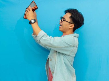 Kot ceketli zavallı Asyalı adam mavi arka planda soyutlanmış, üzgün bir ifadeyle boş bir cüzdan taşıyor..