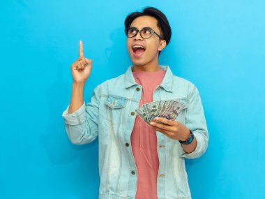 Kot ceketli heyecanlı Asyalı adam elinde para tutuyordu ve mavi arka planda tek bir parmağı havada bir fikir ediniyordu..