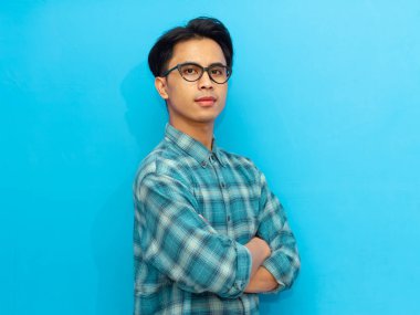 Kollarını kavuşturup kameraya bakan kendine güvenen Asyalı adam. Üniversite, iş ya da profesyonel markalaşma için mükemmel. Mavi arka planda izole edilmiş, reklamlar ya da promosyonlar için ideal..