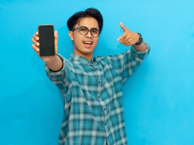 Heyecanlı Asyalı genç adam boş akıllı telefon ekranını işaret ediyor, mavi arka planda izole edilmiş. Mobil bir uygulama, teklif ya da reklam konsepti sunuyorum. Pazarlama ve reklam kullanımı için mükemmel.