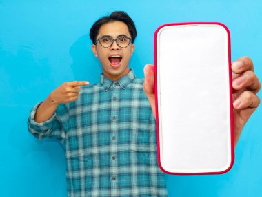 Heyecanlı Asyalı genç adam boş akıllı telefon ekranını işaret ediyor, mavi arka planda izole edilmiş. Mobil bir uygulama, teklif ya da reklam konsepti sunuyorum. Pazarlama ve reklam kullanımı için mükemmel.