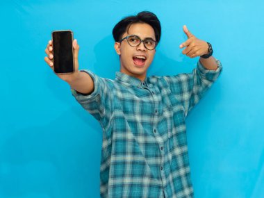 Heyecanlı Asyalı genç adam boş akıllı telefon ekranını işaret ediyor, mavi arka planda izole edilmiş. Mobil bir uygulama, teklif ya da reklam konsepti sunuyorum. Pazarlama ve reklam kullanımı için mükemmel.