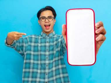 Heyecanlı Asyalı genç adam boş akıllı telefon ekranını işaret ediyor, mavi arka planda izole edilmiş. Mobil bir uygulama, teklif ya da reklam konsepti sunuyorum. Pazarlama ve reklam kullanımı için mükemmel.