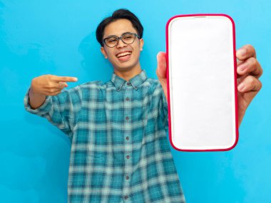 Heyecanlı Asyalı genç adam boş akıllı telefon ekranını işaret ediyor, mavi arka planda izole edilmiş. Mobil bir uygulama, teklif ya da reklam konsepti sunuyorum. Pazarlama ve reklam kullanımı için mükemmel.