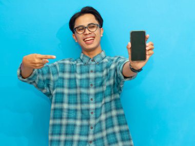 Heyecanlı Asyalı genç adam boş akıllı telefon ekranını işaret ediyor, mavi arka planda izole edilmiş. Mobil bir uygulama, teklif ya da reklam konsepti sunuyorum. Pazarlama ve reklam kullanımı için mükemmel.