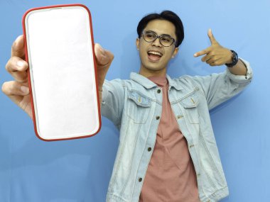 Heyecanlı Asyalı genç adam boş akıllı telefon ekranını işaret ediyor, mavi arka planda izole edilmiş. Mobil bir uygulama, teklif ya da reklam konsepti sunuyorum. Pazarlama ve reklam kullanımı için mükemmel.