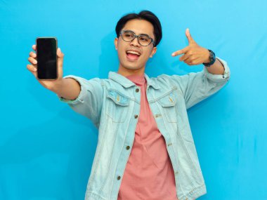 Heyecanlı Asyalı genç adam boş akıllı telefon ekranını işaret ediyor, mavi arka planda izole edilmiş. Mobil bir uygulama, teklif ya da reklam konsepti sunuyorum. Pazarlama ve reklam kullanımı için mükemmel.