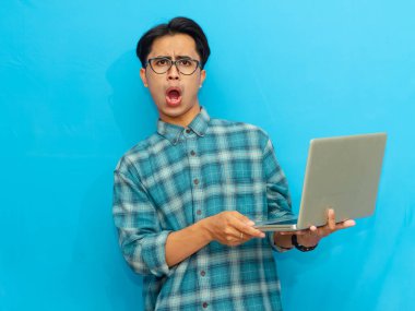 Yakışıklı bir Asyalı erkek öğrenci elinde dizüstü bilgisayarla poz veriyor ve mavi arka planda izole edilmiş komik bir şok ifadesi sergiliyor. Reklam ya da terfi için mükemmel..