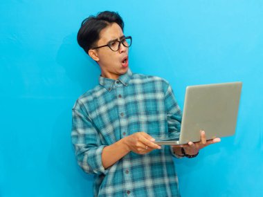 Yakışıklı bir Asyalı erkek öğrenci elinde dizüstü bilgisayarla poz veriyor ve mavi arka planda izole edilmiş komik bir şok ifadesi sergiliyor. Reklam ya da terfi için mükemmel..