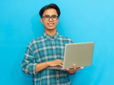Yakışıklı Asyalı genç bir erkek öğrenci gülümsüyor ve dizüstü bilgisayarla poz veriyor, mavi arka planda kameraya bakıyor. Reklam amaçlı ideal..