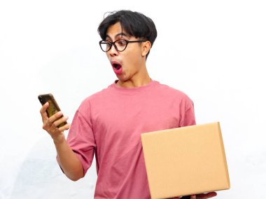 Pembe tişörtlü genç Asyalı bir adam, beyaz bir arka planda izole edilmiş, komik bir ifadeyle cep telefonuna bakarken bir paket kutusu ile poz veriyor..