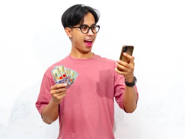 Elinde para tutarak poz veren pembe tişörtlü genç bir Asyalı ve beyaz arka planda izole edilmiş mutlu ve şok edici bir ifadeye sahip bir cep telefonu kullanıyor. Reklam için mükemmel..