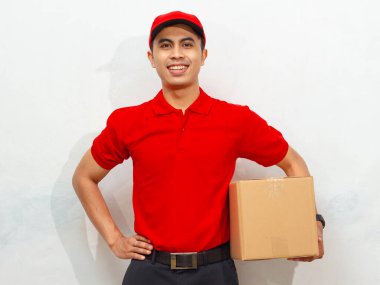 Kırmızı üniformalı ve şapkalı gülümseyen erkek kurye beyaz arka planda izole edilmiş kendinden emin ve dostane bir ifadeye sahip bir paket kutusu tutuyor..