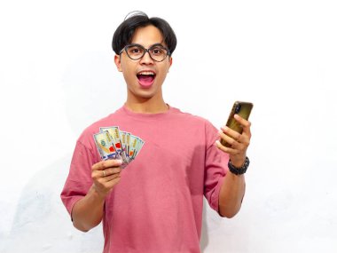 Elinde para tutarak poz veren pembe tişörtlü genç bir Asyalı ve beyaz arka planda izole edilmiş mutlu ve şok edici bir ifadeye sahip bir cep telefonu kullanıyor. Reklam için mükemmel..