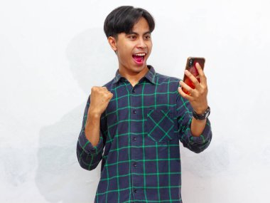 Yakışıklı Asyalı genç bir adamın portresi akıllı bir telefon tutuyor ve yumruğunu sıkıyor ve beyaz bir arkaplanda heyecanlı bir yüz ifadesi sergiliyor. Reklam veya promosyon için mükemmel..