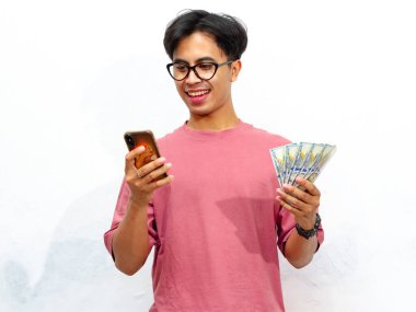 Elinde para tutarak poz veren pembe tişörtlü genç bir Asyalı ve beyaz arka planda izole edilmiş mutlu ve şok edici bir ifadeye sahip bir cep telefonu kullanıyor. Reklam için mükemmel..