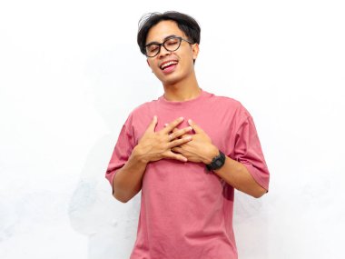 Pembe tişörtlü genç Asyalı bir adamın portresi, göğsünü tutarken rahatlamış bir ifadeyle poz veriyor, beyaz bir arka planda izole edilmiş..