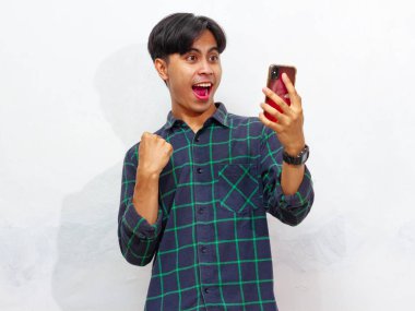 Yakışıklı Asyalı genç bir adamın portresi akıllı bir telefon tutuyor ve yumruğunu sıkıyor ve beyaz bir arkaplanda heyecanlı bir yüz ifadesi sergiliyor. Reklam veya promosyon için mükemmel..