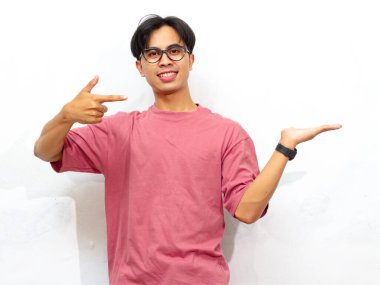 Pembe tişörtlü mutlu genç Asyalı adam beyaz bir arka planda, avucunun içini gösteriyor ve poz veriyor..