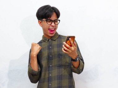 Yakışıklı Asyalı genç bir adamın portresi akıllı bir telefon tutuyor ve yumruğunu sıkıyor ve beyaz bir arkaplanda heyecanlı bir yüz ifadesi sergiliyor. Reklam veya promosyon için mükemmel..