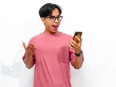 Beyaz arka planda izole edilmiş bir cep telefonu kullanırken mutlu ve şok olmuş bir jest gösteren pembe tişörtlü genç Asyalı bir adamın portresi..