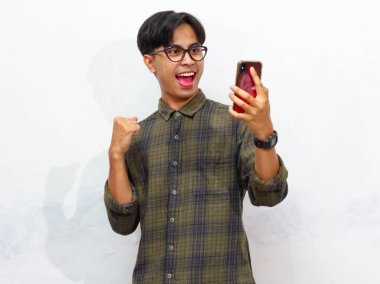 Yakışıklı Asyalı genç bir adamın portresi akıllı bir telefon tutuyor ve yumruğunu sıkıyor ve beyaz bir arkaplanda heyecanlı bir yüz ifadesi sergiliyor. Reklam veya promosyon için mükemmel..